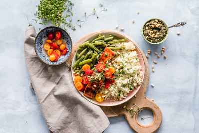 white marble counter with various sized dishes incuding tomatoes green beans cooked grains legumes and a flax napkin on a round wooden board bright colorful plant based meal