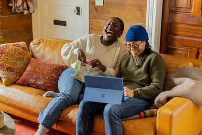 two people sitting on a saddle colored leather couch sofa laughing with a tablet on the lap of one person wearing a blue hat and another person with a big smile head tilted back apparently laughing
