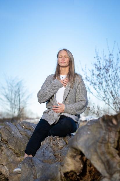 A woman sitting with her hand on her heart and her stomach assessing where her body is in space, and how her body is reacting to it as she invests in a future of better breathing, nervous system regulation and leaving anxiety and body ailments behind.
