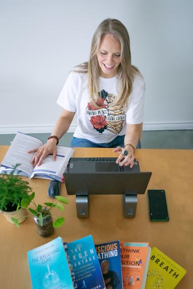 A woman (Randi) sitting in front of a computer for a virtual breathwork session joyfully surrounded by science backed books of how functional breathwork with aid burnt-out, overwhelmed millennial women breathe easier.
