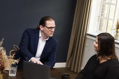 An AI adoption consultant leans over a laptop to discuss strategy with a business professional, illustrating tailored corporate advisory and implementation services.