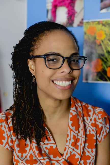 A woman with dark skin and glasses, with dark hair and is smiling and looking directly at the camera.