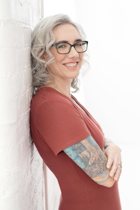 Blaze Schwaller, life coach, leaned against a white brick wall, smiling. She looks at you welcomingly.