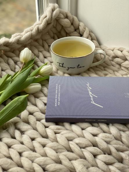 A knitted blanket with cut white tulips laid across it, a journal titled "Purpose" and a cup of tea that says "take your time" sit by a window waiting for you