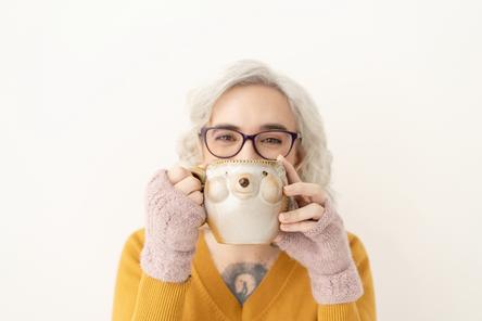 Blaze Schwaller, life coach, holding a mug of tea that looks like a hedgehog. She has smiling eyes and gazes at you from behind the mug. She wears a yellow sweater and knitted pink hand warmers.