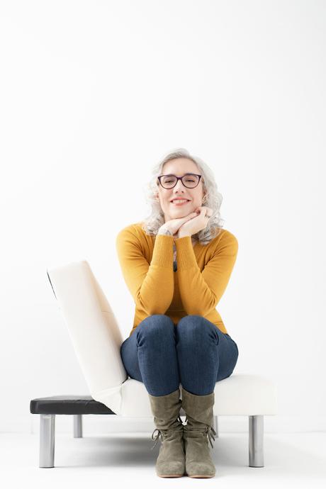 Blaze sits on the edge of a white couch with her hands brought up underneath her chin. She smiles at you warmly.