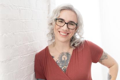 Blaze Schwaller, life coach, smiles at you as she leans against a clean white brick wall. White light floods in. Welcome to your transformation!