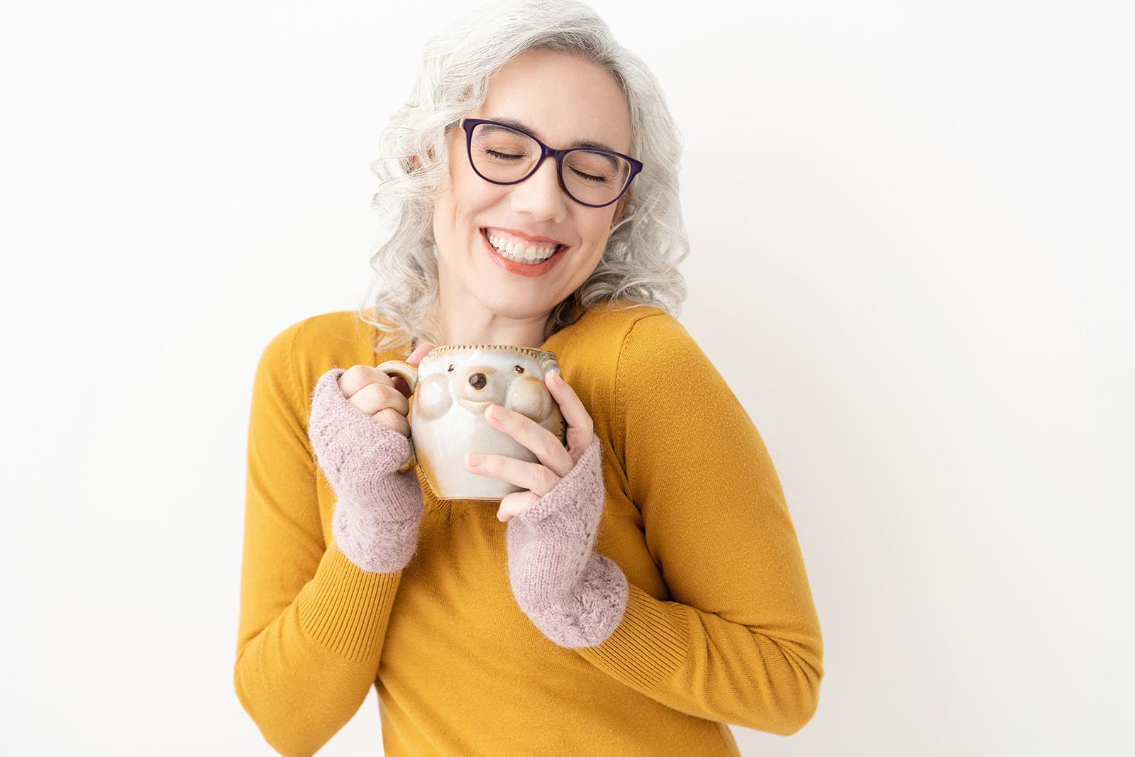 Blaze, delighted, smiling with closed eyes, curly white hair and purple glasses, lifting her hedgehog mug up to her heart, head on view. Joyful, welcoming.