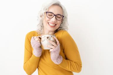 Blaze, delighted, smiling with closed eyes, curly white hair and purple glasses, lifting her hedgehog mug up to her heart, head on view. Joyful, welcoming.