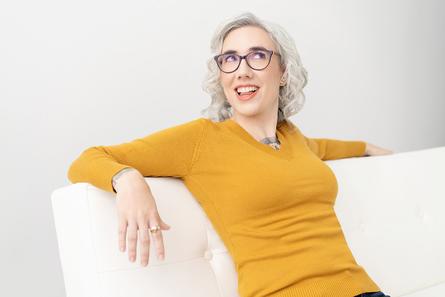 Blaze being silly, leaning back, relaxed on the white couch, looking happy, wearing a yellow sweater and purple glasses.