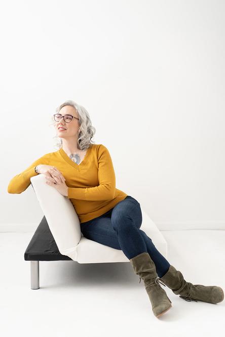 Blaze Schwaller, life coach, sitting sideways on a white couch, smiling toward soft window light.