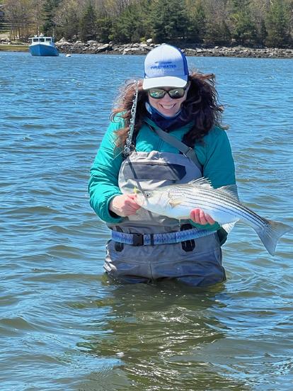 Sarah holding a fish she caught. Wearing waders and a hat and sunglasses, happy