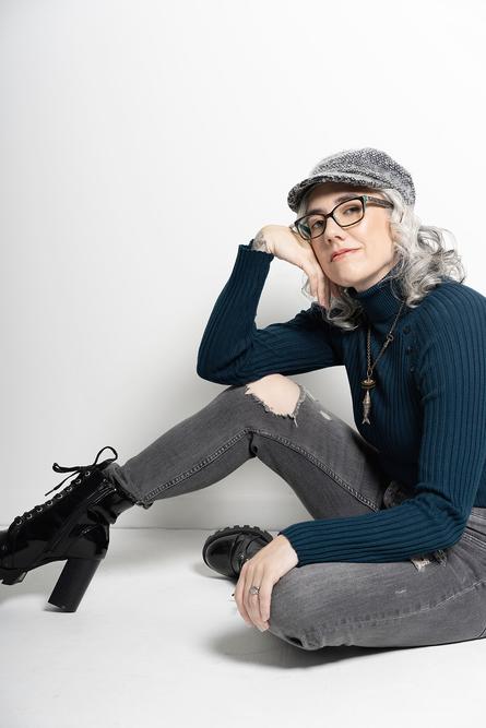 Blaze Schwaller, life coach, sitting on the floor, wearing torn jeans. a blue turtle neck, a grey hat and tall boots, gazing at you with calm seriousness.