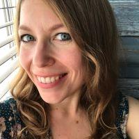 Kirstyn Lazur head shot, smiling in front of a window with white blinds partially opened