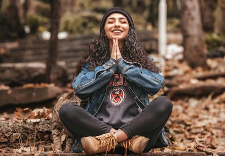 Woman sitting cross-legged in the woods, with her hands together in gratitude, smiling, joyful