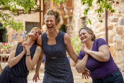 A woman in a blue patterned dress is laughing hard as two coaching colleagues lean into her smiling