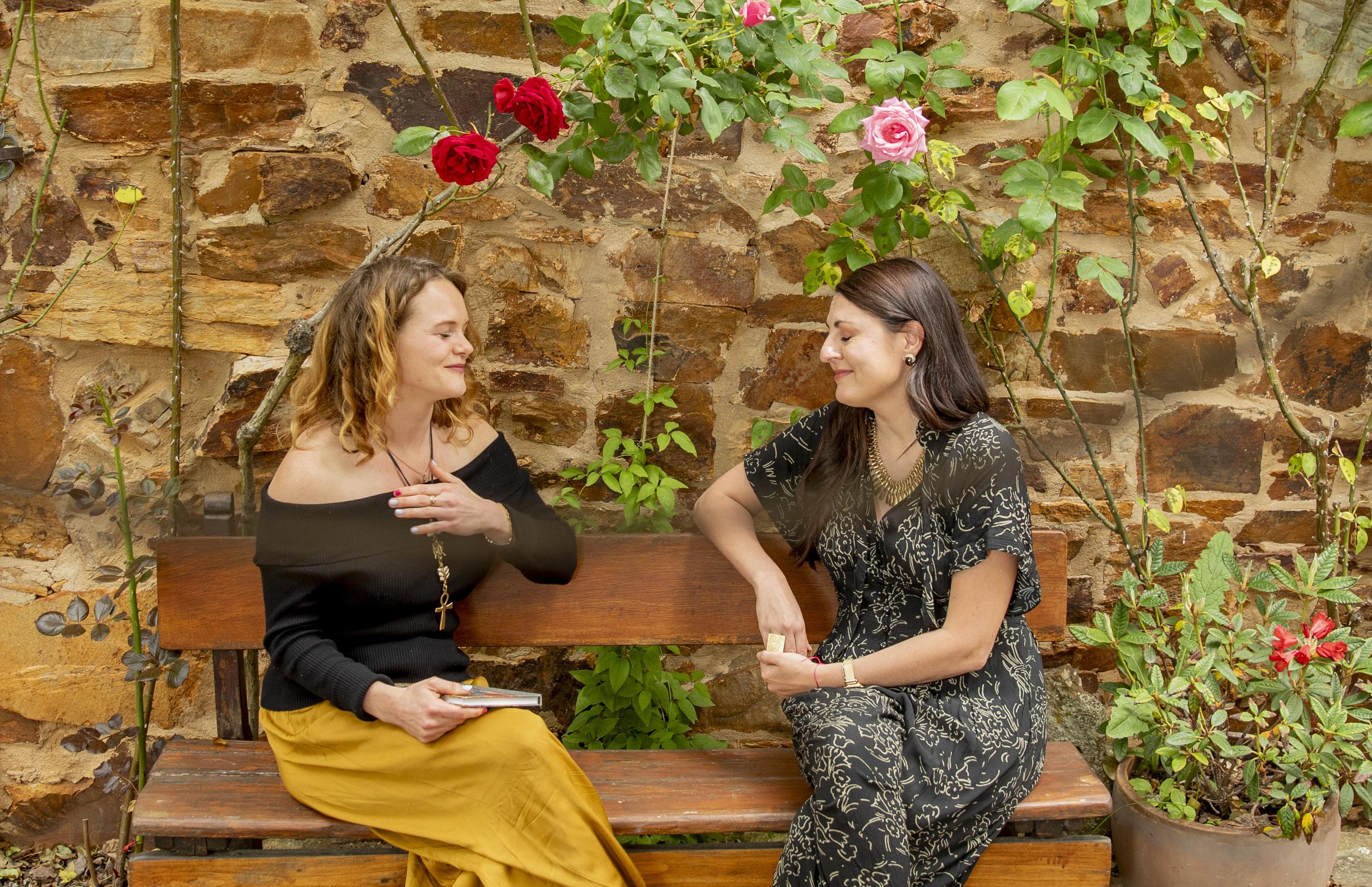 Two women in conversation on a wooden bench in a rose-filled courtyard, embodying the safe, supportive space of Awaken's Transformational Coach Certification training that comes from developing a personal presence practice