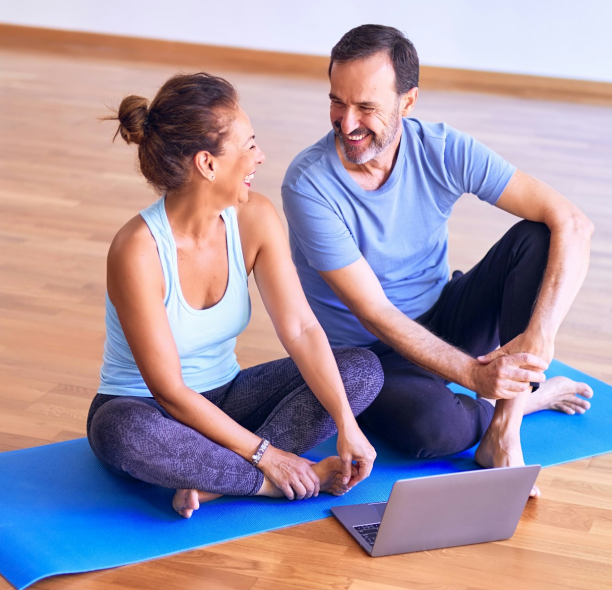 A man and a woman talking to each other, sitting on a yoga mat, laptop open in front of them