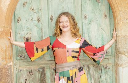A woman in brightly coloured clothes stands in a door way with arms outstretched to the door frame, she is smling