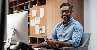a male team leader sitting at his desk in front of his computer smiling and holding his phone