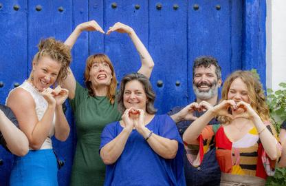 Five coaches stand in front of a blue door in Castrillo de los Polvazres, Spain, each making a heart shape with their hands or arms