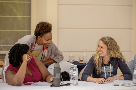 Two women are talking and smiling at each other while seated at a table, with a woman standing leaning over the table. There are water bottles, a glass of water, and a water bottle on the table.