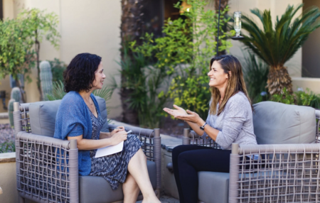 Two women having a conversation outdoors on a patio, sitting on cushioned chairs with greenery in the background.
