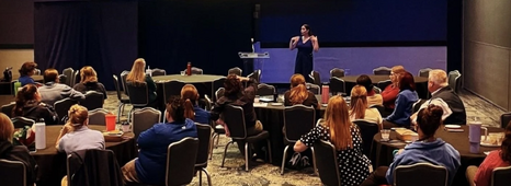 A woman speaks in front of several tables of people facing her