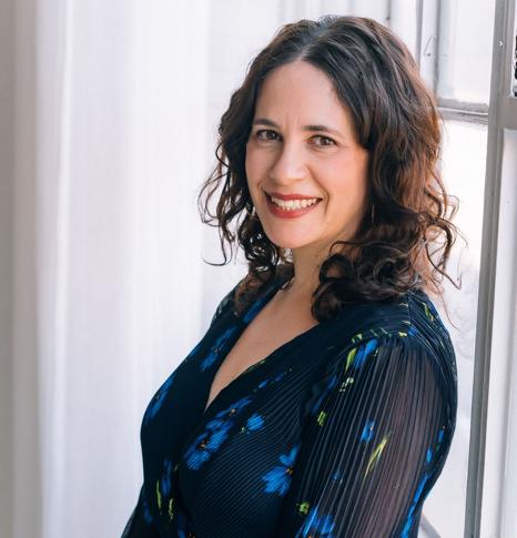 Dr. Jill Wener is smiling and wearing a blue flowered dress as she leans against a window