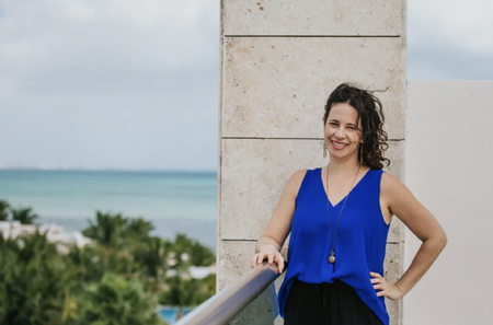 Dr. Jill Wener stands outside on a balcony in a blue shirt, with water and trees in the background