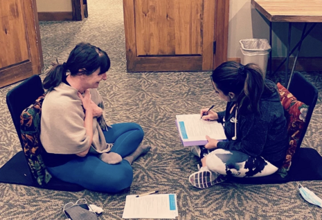 Two women seated in floor chairs laugh as they enjoy participating in a tapping workshop