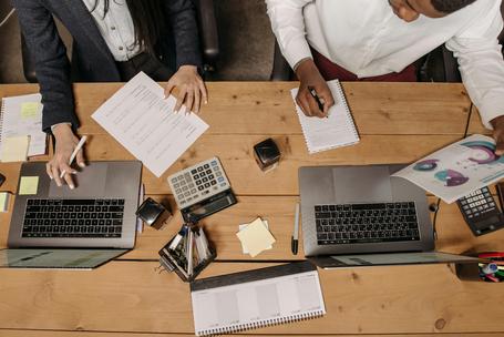 Top-down view of two colleagues sitting at a wooden desk working on laptops, reviewing financial documents, calculators, and taking notes.
