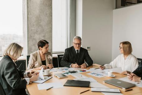 A group of professionals in business attire sitting around a conference table reviewing charts, documents, and reports during a meeting.