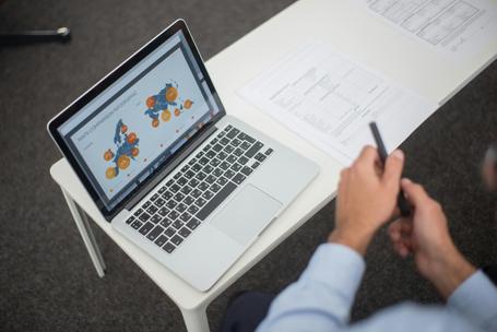 Person holding a pen at a desk while looking at a laptop screen displaying map comparison infographics next to printed paperwork.