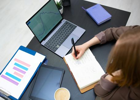 Overhead view of a person writing in a notepad at a desk with a open laptop displaying line graphs, a tablet, and bar chart documents.