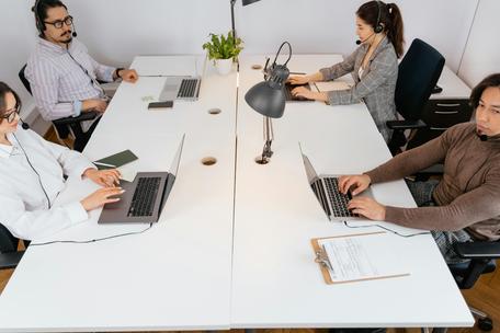 Four office workers wearing headsets sit facing each other at long white desks working on laptops in a modern call center or office setup.