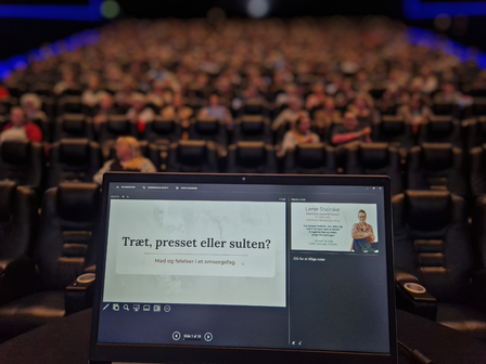 Lene Steinke, diætist og psykoterapeut, computer med præsentation står foran lærred i biograf med fyldt sal af ventende tilhører