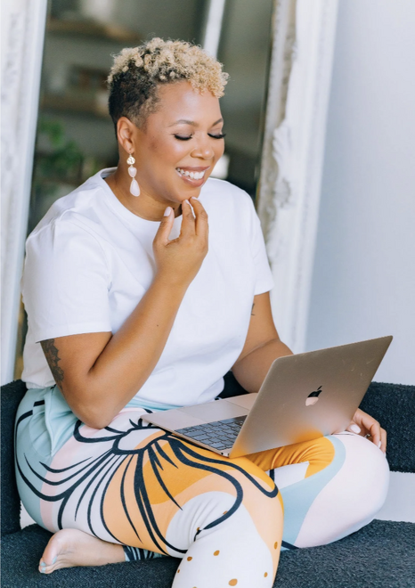 A smiling woman sits with a laptop on her lap as she practices tapping with an online tapping course
