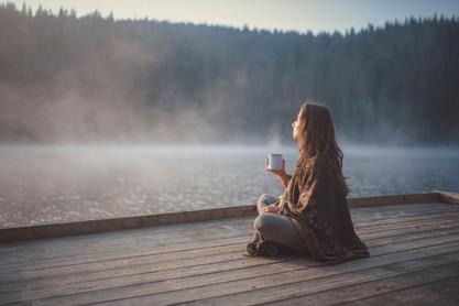 a girl sitting on a dock by a lake swipping a cup of tea and stairing out into the distance