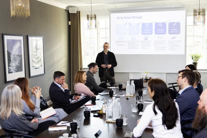 A corporate trainer stands before a boardroom table presenting on a projector screen, leading an AI training workshop on organizational transformation for a group of professionals.