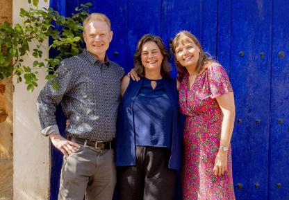 Three of Awaken's coaching faculty stand smiling in front of a deep blue door in dappled sunshine in Spain