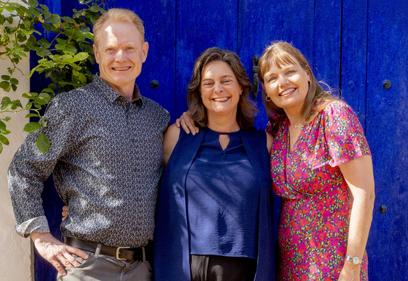 Three of Awaken's coaching faculty stand smiling in front of a deep blue door in dappled sunshine in Spain