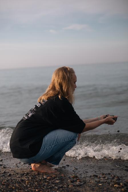A woman squatting by the lake with a handful of sand and stones grateful for all the tools she has acquired in the membership to aid her wanting to live life more bountifully.