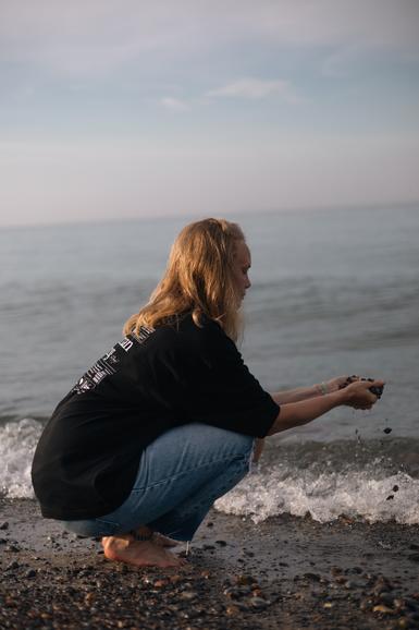 A woman squatting by the lake with a handful of sand and stones grateful for all the tools she has acquired in the membership to aid her wanting to live life more bountifully.