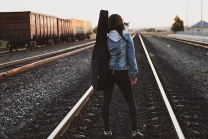 A woman in a jean jacket with a guitar case on her back and akitten on her shoulder walks in the middle of a railroad track