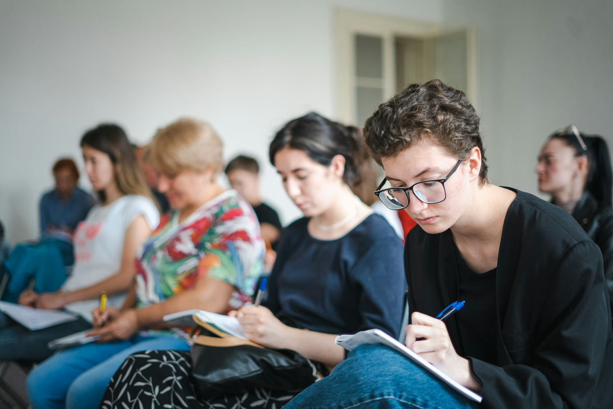 En rekke unge studenter som sitter tett sammen i en sal og skriver i notatblokker på fanget