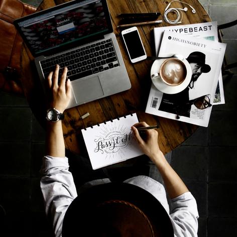 Person using MacBook and writing "Pass it on" in calligraphy on notebook