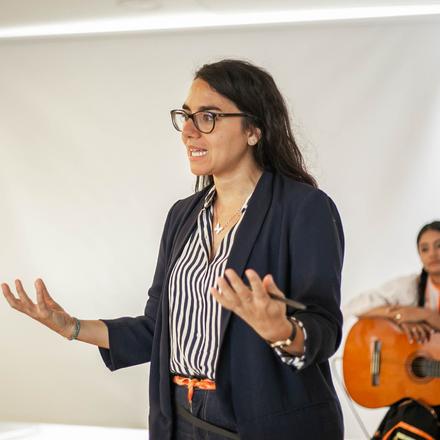 Woman speaking in room with guitarist in backgroud