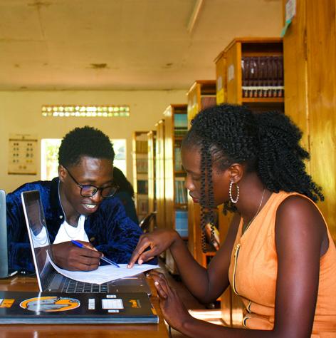A black man and woman writing on paper and using a laptop at a library table