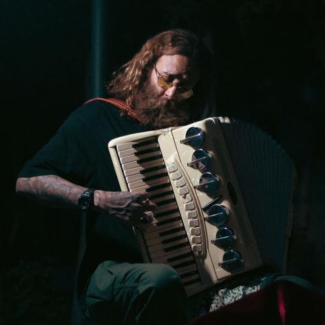A white man with long curly hair playing an accordion